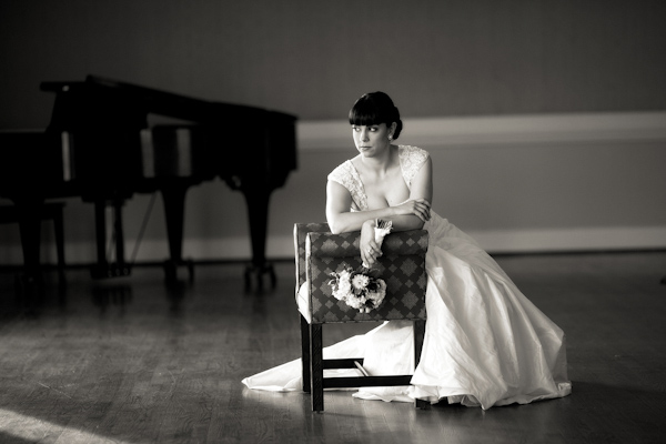 black and white photo - beautiful bride leaning on an antique chair in the middle of an open room with a grand piano in the background - bride is wearing ball gown style dress with retro hairstyle - photo by North Carolina based wedding photographers Cunningham Photo Artists